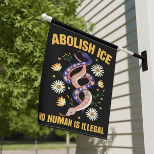 May include: Black flag with the text "ABOLISH ICE" in yellow, and "NO HUMAN IS ILLEGAL" in yellow. The flag features a colorful snake design with flowers and leaves.
