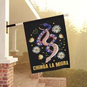 May include: Black flag featuring a stylized snake illustration in blue, pink, and white, surrounded by white daisies and yellow flowers. The text "CHINGA LA MIGRA" is in yellow. The flag is hanging outside.