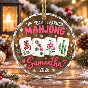 Può includere: Un ornamento circolare in acrilico trasparente con il testo THE YEAR I LEARNED MAHJONG, Samantha e 2026. Il design include quattro tessere del mahjong con simboli rossi e verdi, circondate da piccoli fiocchi di neve bianchi, bacche rosse e cuori rosa.