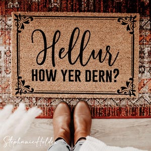 Könnte beinhalten: Rechteckige Fußmatte aus Kokosfaser mit dem schwarzen Schriftzug "Hellur HOW YER DERN?". Die Matte hat dekorative schwarze Eckakzente. Braune Stiefel sind am unteren Bildrand zu sehen, auf einem Teppich stehend.