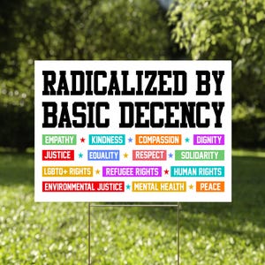Könnte beinhalten: Ein weißes Schild mit dem Text "RADICALIZED BY BASIC DECENCY" in fetter schwarzer Schrift. Darunter listen farbige Rechtecke Werte wie "EMPATHY", "KINDNESS" und "EQUALITY" auf. Das Schild steht in einem grasbewachsenen Garten mit verschwommenem grünem Hintergrund.
