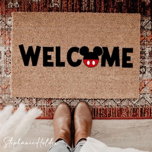 Può includere: Un zerbino rettangolare con la scritta "WELCOME" in grandi lettere nere. La "O" è sostituita dalla testa di Topolino, con orecchie nere e un viso rosso e bianco. Il tappeto è di colore marrone chiaro.