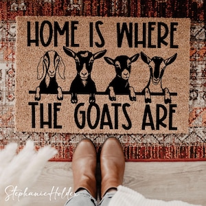 Könnte beinhalten: Eine rechteckige Fußmatte mit dem Text "HOME IS WHERE THE GOATS ARE" in Schwarz. Die Matte zeigt vier schwarze Ziegenillustrationen. Die Matte ist braun und liegt auf einem gemusterten Teppich.