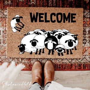May include: A rectangular doormat with the word "WELCOME" in bold black letters. The mat features a cartoon illustration of a flock of sheep, with one sheep jumping. The mat is brown and placed on a patterned rug.