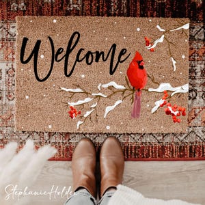 May include: A rectangular doormat with the word "Welcome" in black script. A red cardinal bird perches on a snow-covered branch with red berries. The mat is brown with white speckles, and the background includes a patterned rug and brown boots.