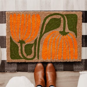 May include: A rectangular doormat with a floral design. The mat features two large orange flowers with green stems and leaves against a tan background. The mat is placed on a patterned rug, with brown boots visible at the bottom.