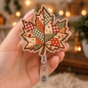 May include: A maple leaf-shaped badge reel featuring a patchwork design with various fabric patterns including red and white gingham, green plaid, and floral prints in orange and cream, finished with dark brown stitched edges and a clear plastic clip.
