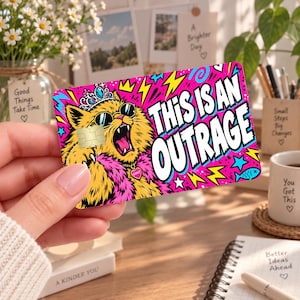 May include: A rectangular credit card skin featuring a vibrant pop art illustration of a yellow cat wearing a blue crown, sunglasses, and a pink boa. The text THIS IS AN OUTRAGE is printed in bold white letters against a bright pink background.