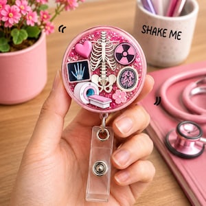 Op de afbeelding: Een ronde roze badgehouder gevuld met kleine roze kraaltjes en medische bedeltjes, waaronder een menselijke ribbenkast, een hart, een brein, een röntgenfoto van een hand, een stralingssymbool en een MRI-scanner met een doorzichtig bandje.
