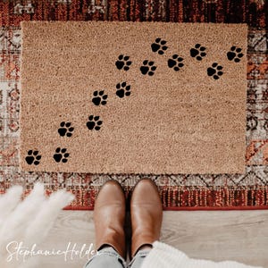 Puede incluir: Felpudo rectangular de material marrón claro y texturizado. Huellas de patas negras dispuestas en diagonal sobre la alfombra. El felpudo está colocado sobre una alfombra estampada y un suelo de madera.