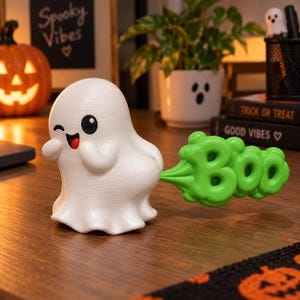 May include: A white ghost figurine with a winking face, a red tongue, and a green cloud-shaped speech bubble attached to its back that features the word Boo in raised, rounded letters. The ghost has a smooth, matte finish and a rounded shape.