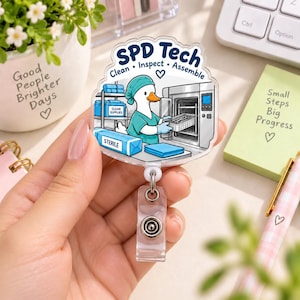Può includere: Un porta badge in acrilico trasparente con un'anatra cartoon in divisa medica color ottanio e cuffia chirurgica, che inserisce un vassoio in un'autoclave. Il testo riporta SPD Tech, Clean, Inspect, Assemble e Sterile.