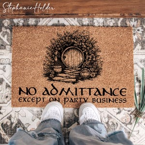 May include: A rectangular coir doormat featuring a black illustration of a round door framed by plants. The text "NO ADMITTANCE except ON PARTY BUSINESS" is below. The mat is on a patterned rug, with feet in white trainers.