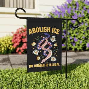 May include: Black garden flag with the text "ABOLISH ICE" and "NO HUMAN IS ILLEGAL" in yellow. The flag features a colorful illustration of a snake, flowers, and leaves. The flag is displayed on a black metal stand in a grassy yard.