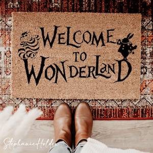 Peut inclure: Un paillasson rectangulaire avec les mots "Welcome to Wonderland" en noir. Le tapis présente des illustrations du Chat du Cheshire et du Lapin Blanc. Des bottes marron sont visibles en bas.