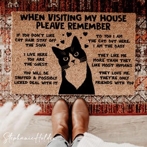 Könnte beinhalten: Eine rechteckige Fußmatte mit einer Katzenillustration und humorvollem Text. Die Matte ist beige mit schwarzem Text und einem schwarz-weißen Katzendesign. Der Text lautet "When visiting my house, please remember..." und andere katzenbezogene Sätze.