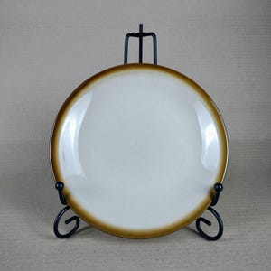 Pode incluir: Um prato redondo de cerâmica branca com borda marrom, exibido em um suporte de metal preto. O prato tem um acabamento brilhante e um design simples. O suporte tem uma base decorativa com volutas.