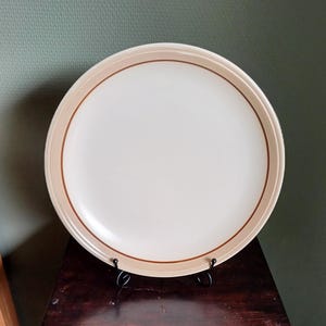 Op de afbeelding: Een grote, ronde, crèmekleurige keramische dinerbord met een bruine rand en een dunne bruine lijn rond de rand.