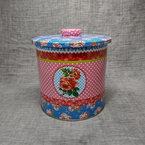 Könnte beinhalten: Eine runde, rosafarbene und blaue Metalldose mit einem Blumendesign. Die Dose hat einen weißen Deckel mit rosa Punkten. Das Blumendesign zeigt rosa Rosen und grüne Blätter.
