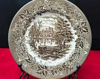 May include: A round ceramic plate featuring a brown and white transferware scene of a person in a small boat on a river near a stone building. The rim is decorated with a detailed border of leaves, berries, and small flowers in brown tones.