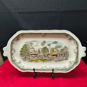 May include: An oblong ceramic serving tray with handles on each side, featuring a transferware illustration of a village scene with houses, trees, and a church. The border has a decorative brown lattice pattern on a cream-coloured background.