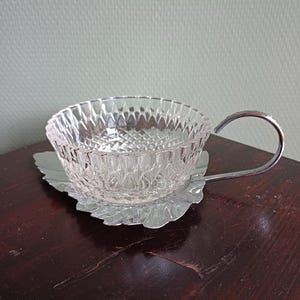May include: A clear glass teacup with a diamond pattern sits on a silver leaf-shaped saucer. The cup has a curved silver handle. The teacup and saucer are on a dark brown wooden surface.