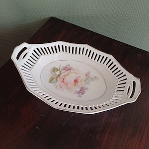 Könnte beinhalten: Weiße ovale Servierplatte mit einem zarten Blumendesign mit einer rosa Rose und lila Blüten. Die Platte hat einen dekorativen durchbrochenen Rand und zwei Griffe.