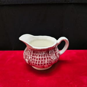 May include: A small, white ceramic creamer with a handle and spout. The creamer features a burgundy-colored illustration of a building with the text "COACH OF!". The creamer sits on a red surface against a black background.