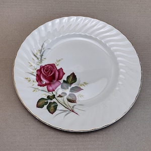 Può includere: Un piatto da portata in ceramica bianca con un bordo dorato presenta una singola rosa rosa con foglie e steli verdi su uno sfondo bianco.