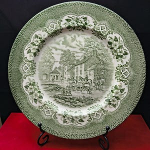 Op de afbeelding: Een ronde keramische bord met een groen en wit transferpatroon. Het midden toont een landelijk tafereel met stenen gebouwen, paarden en ruiters. De brede rand is versierd met ingewikkelde bloemmotieven en rasterpatronen.