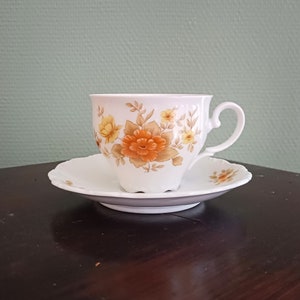 Può includere: Una tazza da tè e un piattino bianchi con un motivo floreale in tonalità arancio, giallo e marrone. La tazza ha un manico delicato e il piattino ha un bordo ondulato.