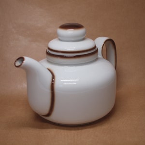 Könnte beinhalten: Weiße Teekanne aus Keramik mit braunem Deckel und braunen Streifenakzenten. Die Teekanne hat einen runden Körper und einen Ausguss auf der linken Seite.