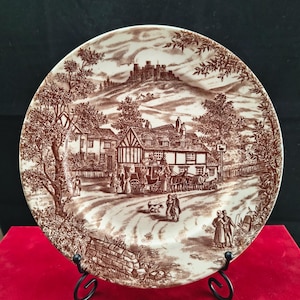 Op de afbeelding: Bruin en wit decoratief bord met een gedetailleerde illustratie van een landelijke scène, inclusief een kasteel, gebouwen en een paardenkoets. Het bord heeft een vintage esthetiek. Het bord wordt gepresenteerd op een zwarte metalen standaard.