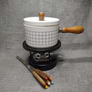 Op de afbeelding: Witte keramische fonduepan met een blauw-wit geruit patroon en een houten handvat. De pan staat op een zwarte standaard met een zilveren brander. Er zijn vijf houten fonduevorken met gekleurde uiteinden.