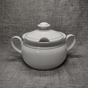 May include: White porcelain soup tureen with a lid and two handles. The tureen has a black trim around the rim and a black line around the body.