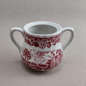 Może przedstawiać: Biały ceramiczny dzbanek do śmietanki z czerwonym wzorem kwiatowym. Dzbanek ma uchwyt po obu stronach i dzióbek. Wzór przedstawia domek, drzewa i chmury.