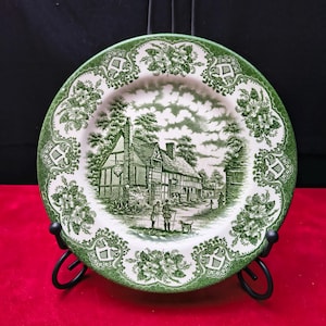 Op de afbeelding: Een ronde keramische bord met een groen en wit transferware patroon. Het midden toont een vakwerkhuis met twee figuren en een hond. De brede rand is versierd met herhalende bloemmotieven en krulwerkontwerpen.
