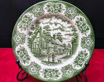 May include: A round ceramic plate featuring a green and white transferware pattern. The center depicts a half-timbered building with two figures and a dog. The wide border is decorated with repeating floral motifs and scrollwork designs.