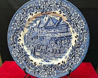 May include: A round ceramic plate featuring a blue and white transferware pattern. The center depicts a historical street scene with timber-framed buildings, a horse-drawn cart, and figures. The rim is decorated with a dense floral scroll motif.