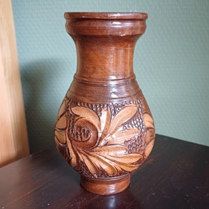 May include: A brown wooden vase with intricate carved floral designs. The vase has a wide base and a narrow neck.