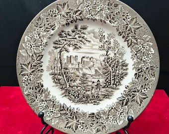May include: A round ceramic plate featuring a sepia-toned transferware pattern. The center depicts a landscape with a large stone castle, trees, and two figures. The wide border is decorated with a detailed relief of grape leaves and clusters.