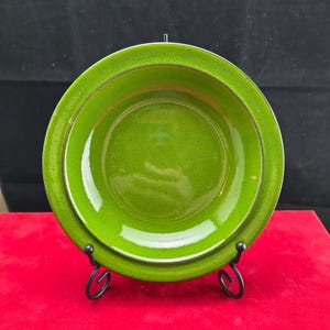 Op de afbeelding: Een heldergroene keramische plaat met een gespikkelde textuur, gepresenteerd op een zwarte metalen standaard. De plaat heeft een brede rand met een contrasterende bruine en witte rand. De plaat is rond en lijkt een serveerschaal te zijn.