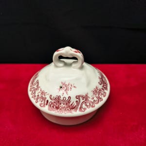 Può includere: Un coperchio in ceramica bianca caratterizzato da un motivo decorativo floreale e a volute color bordeaux lungo il bordo e da una piccola maniglia ad anello sulla parte superiore. Il coperchio ha una finitura liscia e lucida con un foro.