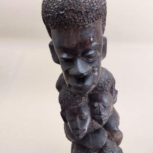 May include: A dark brown wooden sculpture featuring multiple carved faces. The sculpture has a textured surface and detailed facial features, including eyes, noses, and mouths. The top of the sculpture has a detailed carved pattern resembling hair.