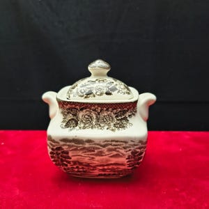 May include: A white ceramic sugar bowl with a lid, featuring a brown floral and landscape pattern. The bowl has two small handles and a decorative knob on the lid. The design is reminiscent of vintage tableware.