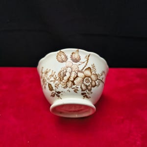 May include: A small, off-white ceramic bowl featuring a brown floral transferware pattern of roses and leaves. The bowl has a scalloped rim and a pedestal base. The exterior surface is decorated with detailed botanical illustrations in brown ink.