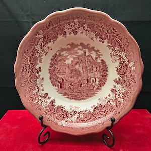 Puede incluir: Un cuenco de cerámica redondo con borde festoneado, que presenta un patrón de transferencia de color rosa oscuro sobre un fondo crema. El centro representa una escena de edificio histórico con figuras, mientras que el borde está decorado con un motivo floral.