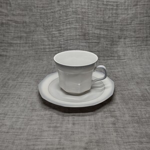 May include: A white porcelain teacup and saucer set. The cup has a rounded shape and a handle. The saucer is round with a slightly raised edge.