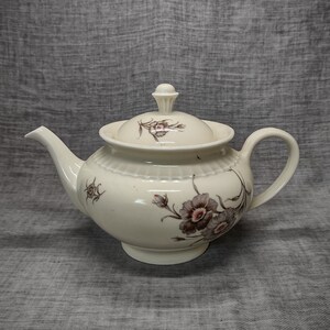 Puede incluir: Una tetera de cerámica blanca con un diseño floral. La tetera tiene una tapa y un asa. Las flores son rosas y marrones.
