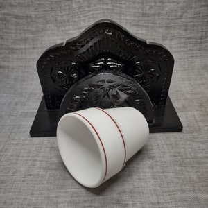 May include: A black wooden cup holder with intricate carvings. The holder has a white ceramic cup with a red band around the top.